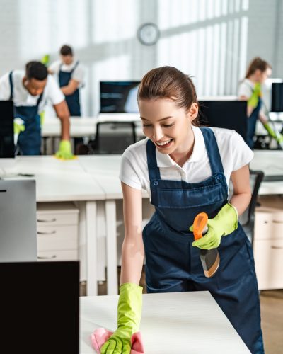 cheerful cleaner in overalls cleaning office desk with rag